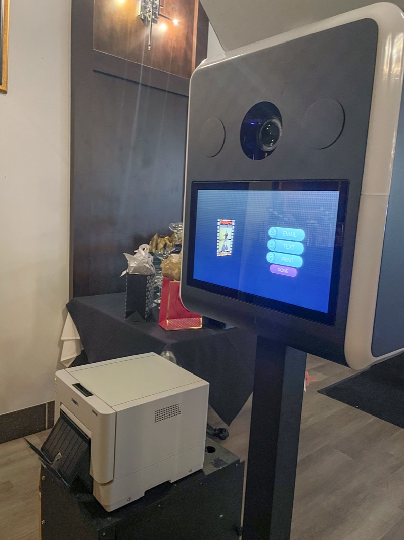 PBMV photo booth: a white kiosk with a camera and touchscreen offering email, text and print options, with a compact photo printer beside it