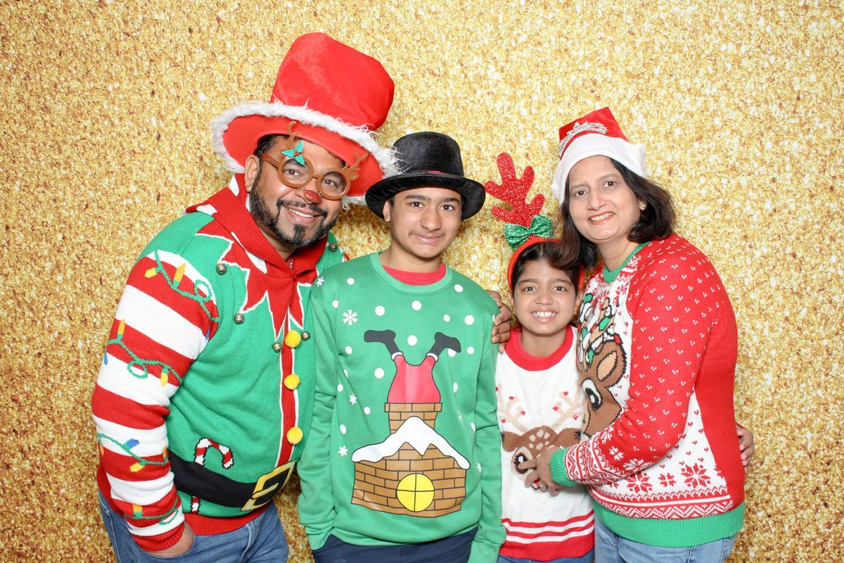 A family in holiday sweaters, Santa hats and reindeer antlers in front of a gold glitter backdrop