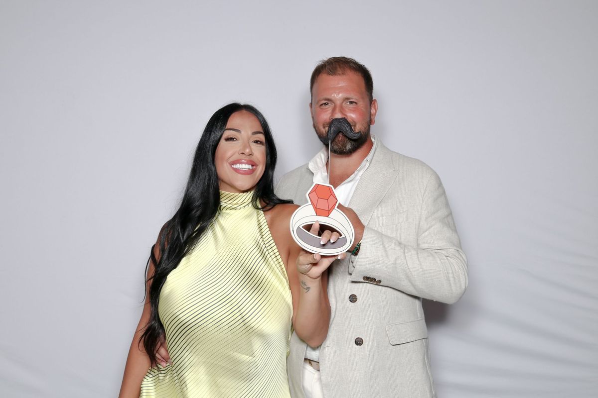 A couple posing with a giant ring prop and a moustache on a stick in front of a white backdrop