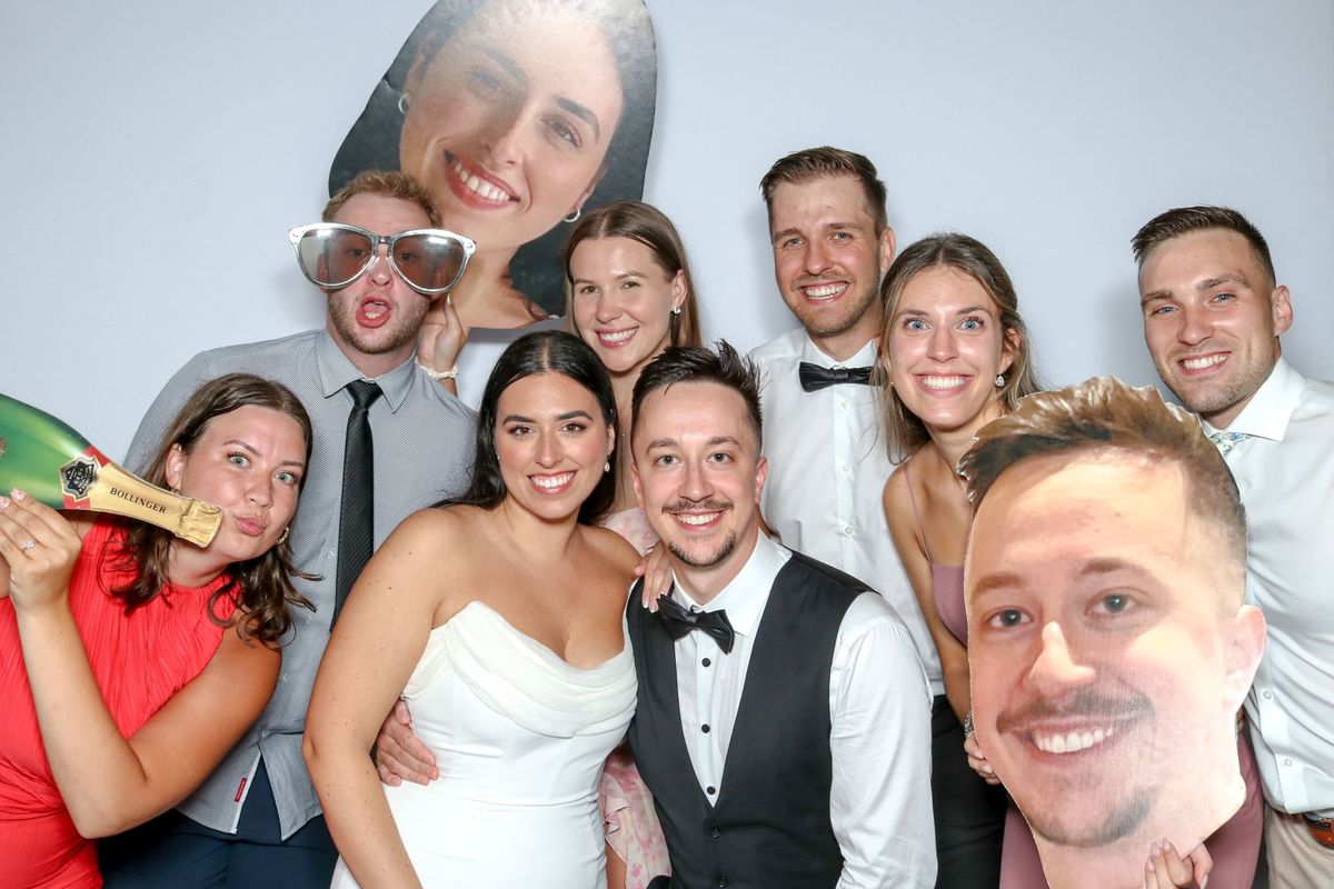 A bride and groom surrounded by wedding guests with giant face cut-outs, oversized sunglasses and a champagne bottle prop