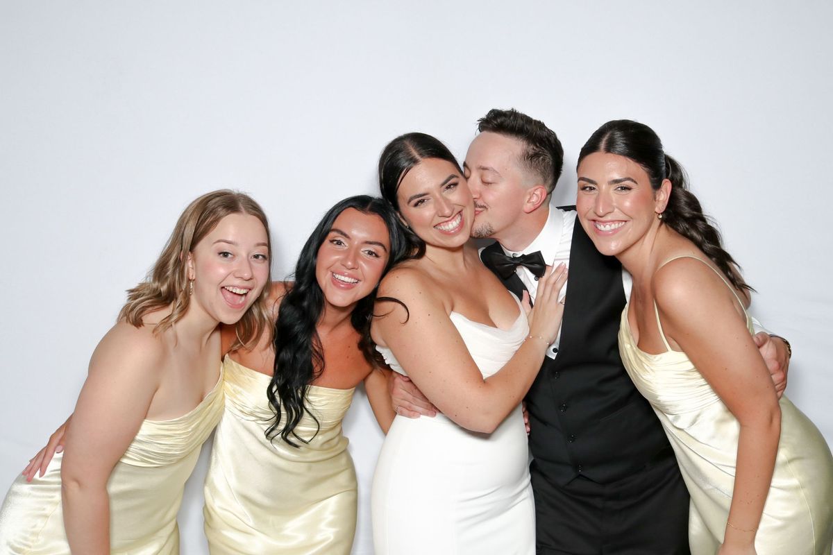A bride and groom with three bridesmaids in pale yellow dresses, laughing in front of a white backdrop as the groom kisses the bride’s cheek