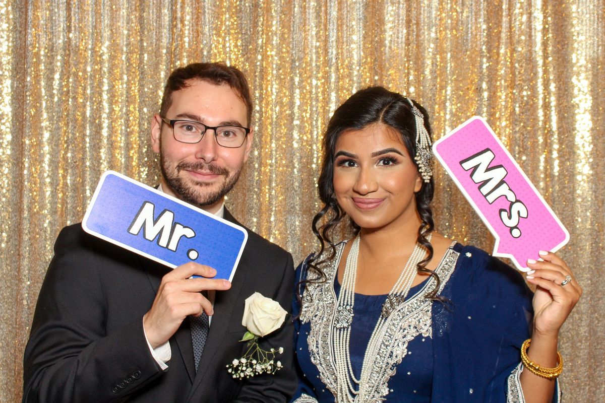 A newlywed couple holding “Mr.” and “Mrs.” signs in front of a gold sequin backdrop