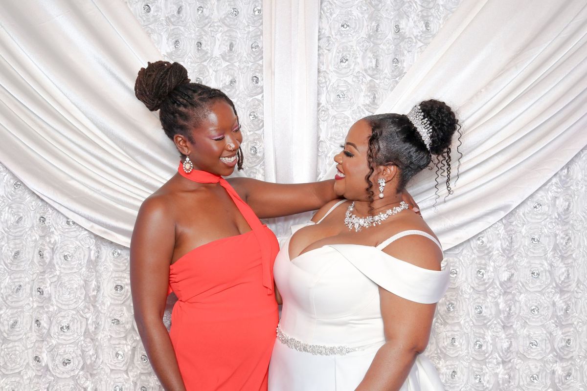 A bride and a friend smiling at each other in front of a white rosette and drape backdrop